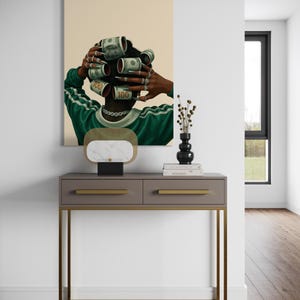 May include: A framed artwork depicts a person with dollar bills rolled into curlers. The person wears a green and white striped shirt and a silver chain. A gray console table with gold legs and a marble lamp are in front of the artwork.