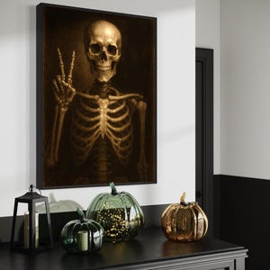 Puede incluir: Una obra de arte enmarcada representa un esqueleto dorado haciendo el signo de la paz. Debajo, un mueble negro exhibe calabazas decorativas de vidrio en verde y dorado, y una linterna negra con una vela. La escena evoca un tema de Halloween.