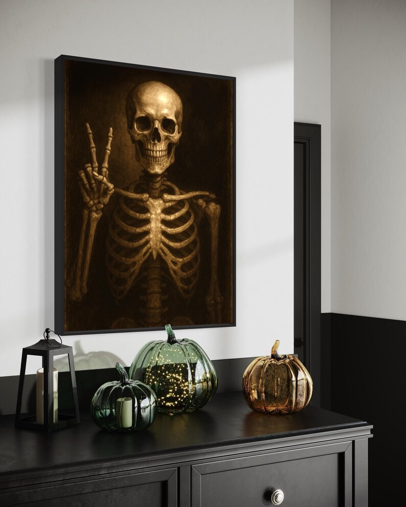 Puede incluir: Una obra de arte enmarcada representa un esqueleto dorado haciendo el signo de la paz. Debajo, un mueble negro exhibe calabazas decorativas de vidrio en verde y dorado, y una linterna negra con una vela. La escena evoca un tema de Halloween.
