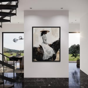 May include: A framed artwork showing a person on a black horse, wearing a white dress and a wide-brimmed hat. The background has neutral tones. The artwork is displayed on a white wall in a modern interior, with a black marble floor.