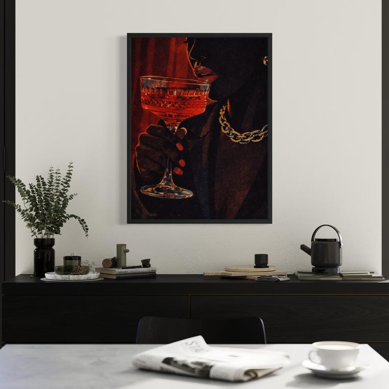 May include: A framed art print depicts a person holding a glass of red liquid. The person is wearing a gold chain necklace and a dark blazer. The background is a deep red color.