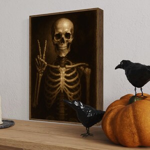 Puede incluir: Una obra de arte enmarcada representa un esqueleto dorado haciendo el signo de la paz. Dos figuras de cuervos negros y una calabaza naranja se encuentran sobre una superficie de madera. Tres velas blancas est&aacute;n en una bandeja plateada. Decoraci&oacute;n de Halloween.