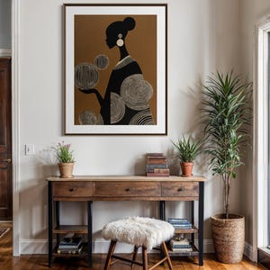 May include: A framed art print featuring a black silhouette of a woman holding orbs with intricate white line details. The artwork is set against a brown background and framed in a dark wood. The print hangs above a wooden desk.