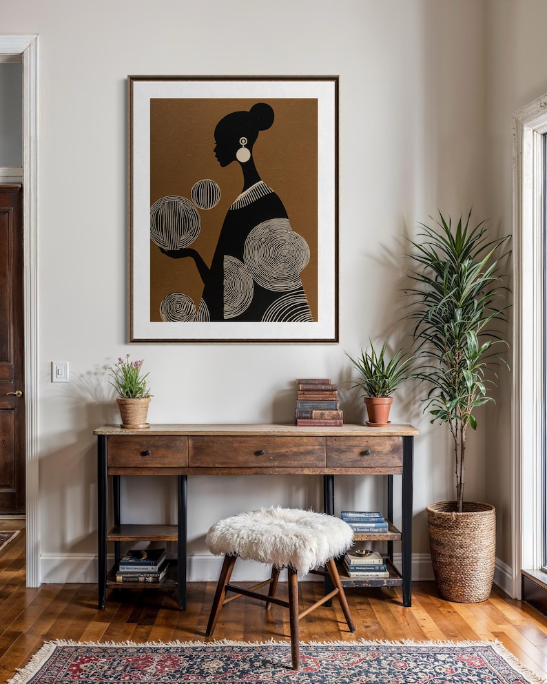 May include: A framed art print featuring a black silhouette of a woman holding orbs with intricate white line details. The artwork is set against a brown background and framed in a dark wood. The print hangs above a wooden desk.