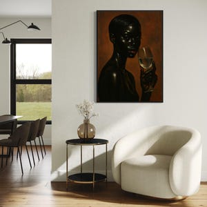 May include: A framed portrait of a person with dark skin holding a wine glass. The artwork is displayed on a white wall, with a small side table and a cream-colored armchair in the foreground. The room has hardwood floors and a large window.