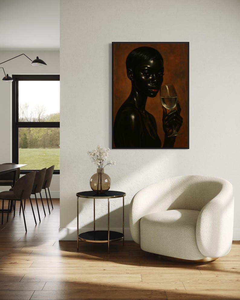 May include: A framed portrait of a person with dark skin holding a wine glass. The artwork is displayed on a white wall, with a small side table and a cream-colored armchair in the foreground. The room has hardwood floors and a large window.
