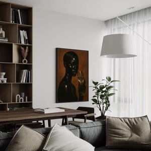 May include: An interior shot featuring a framed artwork of a person holding a glass of wine. The artwork hangs on a white wall above a gray sofa with decorative pillows. A wooden bookshelf and table are also visible.