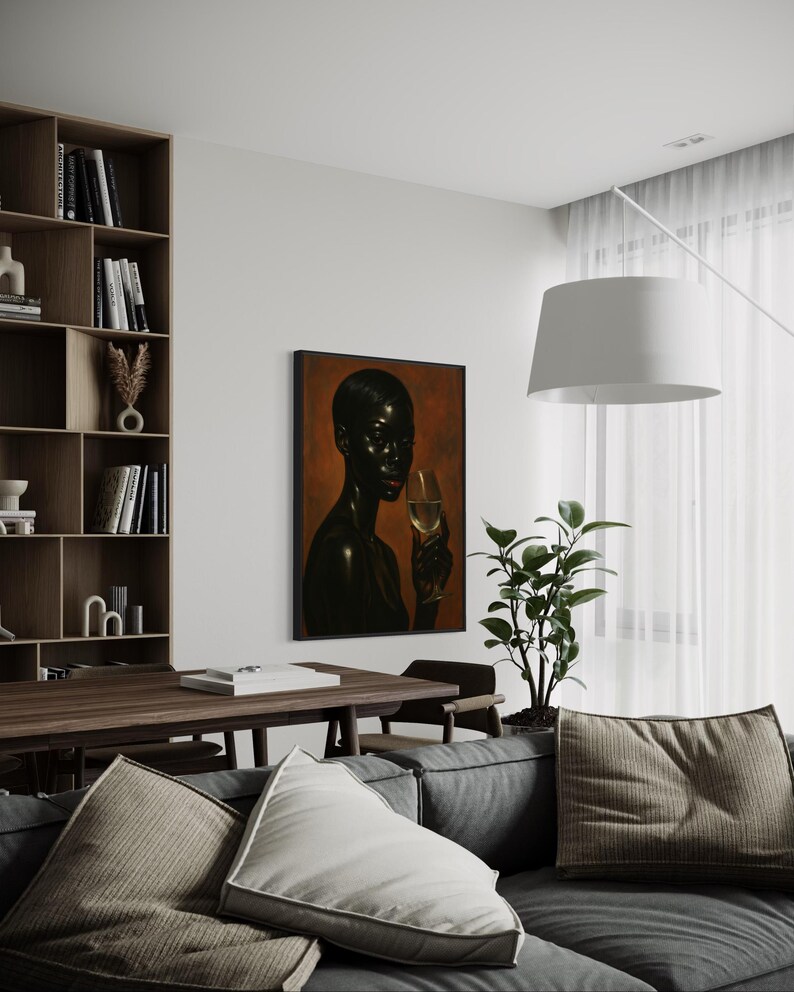 May include: An interior shot featuring a framed artwork of a person holding a glass of wine. The artwork hangs on a white wall above a gray sofa with decorative pillows. A wooden bookshelf and table are also visible.