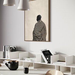 May include: A framed artwork shows a person in a striped garment, set against a neutral background. The room features white shelves with books, a black teapot, a cup, and a round white table. Two white pendant lights hang from the ceiling.
