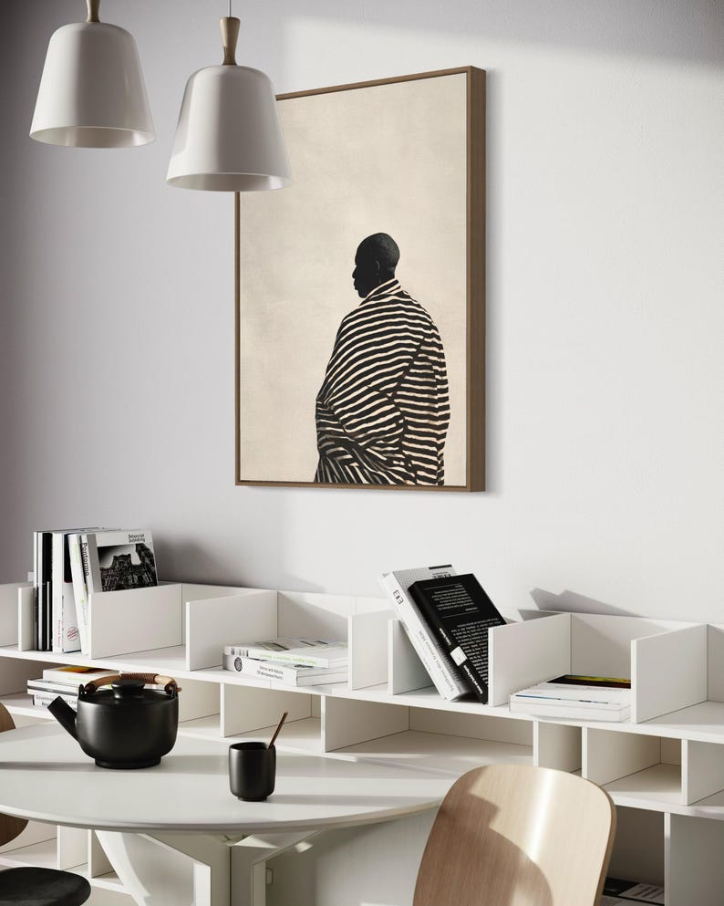 May include: A framed artwork shows a person in a striped garment, set against a neutral background. The room features white shelves with books, a black teapot, a cup, and a round white table. Two white pendant lights hang from the ceiling.