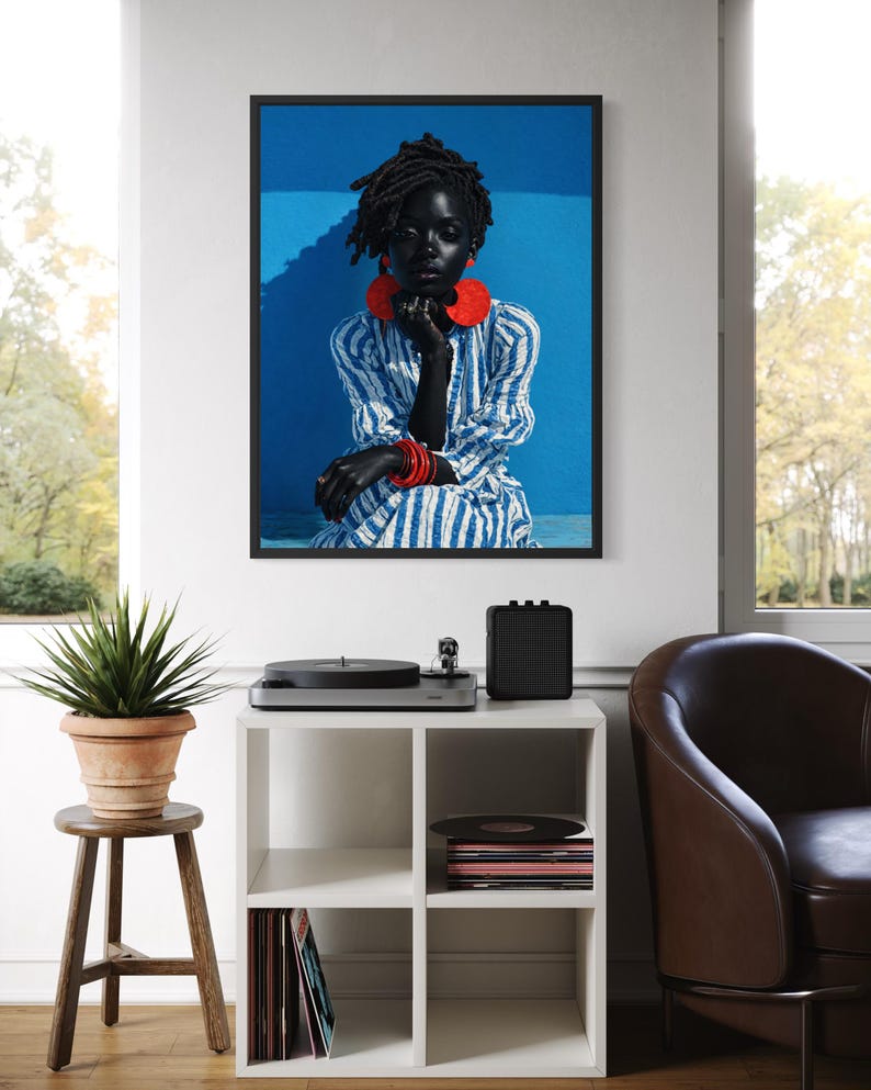 May include: A framed portrait of a person with dark skin, wearing a blue and white striped dress and red jewellery. The portrait hangs above a white cube shelf with a record player, speaker, and vinyl records. A potted plant sits on a wooden stool.