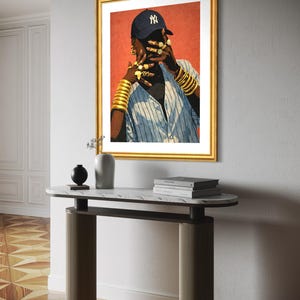 May include: A framed art print featuring a person wearing a navy blue baseball cap with a white NY logo, gold jewelry, and a blue and white striped shirt. The artwork is displayed above a marble-topped console table.