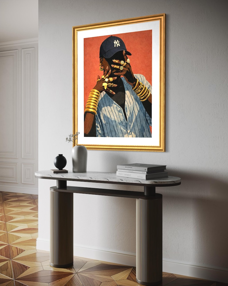 May include: A framed art print featuring a person wearing a navy blue baseball cap with a white NY logo, gold jewelry, and a blue and white striped shirt. The artwork is displayed above a marble-topped console table.