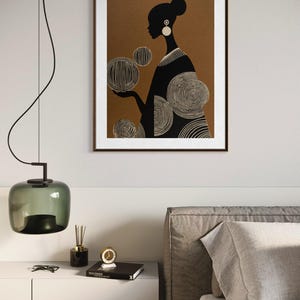 May include: A framed art print featuring a silhouette of a person with a bun, wearing earrings, and a patterned dress. The figure is set against a brown background with white circular designs. The print is in a brown frame.