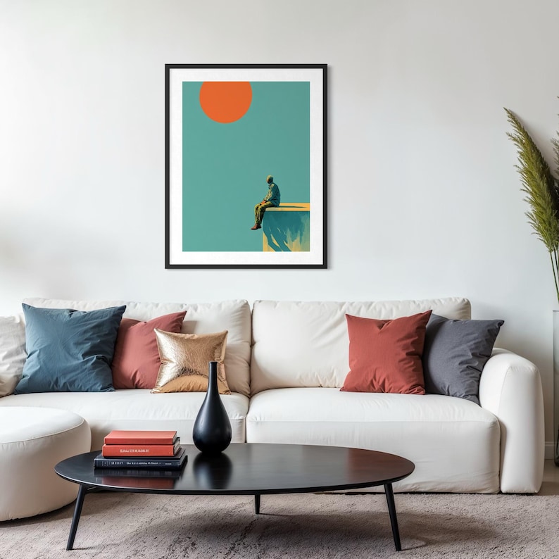 May include: Framed art print featuring a person sitting on a ledge under a large orange sun against a teal sky. The artwork has a minimalist style and is displayed above a white sofa with colorful throw pillows.