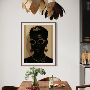 May include: A framed art print featuring a stylized portrait of a woman with dark skin, wearing gold-rimmed sunglasses, a pearl necklace, and ornate earrings. The artwork is set against a neutral background, with a brown wooden frame. The image is displayed in a dining room setting.