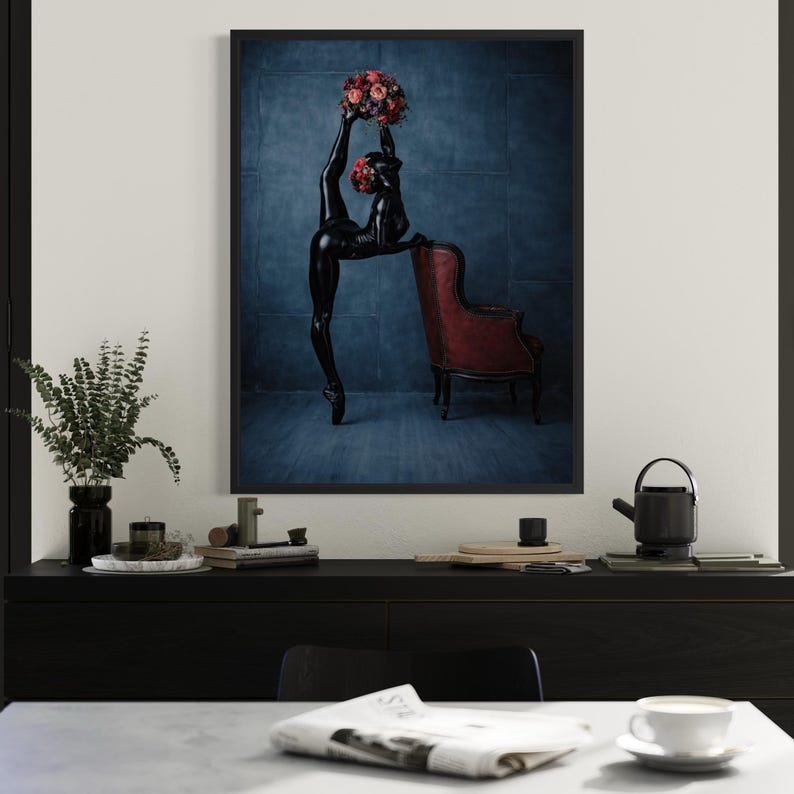 May include: A framed art print showing a black figure in a ballet pose with two flower bouquets. The figure leans on a red armchair against a blue backdrop. The artwork is displayed above a black cabinet.