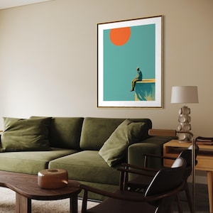 May include: Framed art print featuring a person sitting on a ledge, with an orange sun against a teal sky. The artwork is in a gold frame. The room also includes a green sofa, wooden coffee table, and a lamp.
