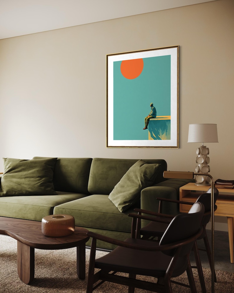 May include: Framed art print featuring a person sitting on a ledge, with an orange sun against a teal sky. The artwork is in a gold frame. The room also includes a green sofa, wooden coffee table, and a lamp.