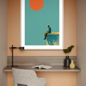May include: Framed art print featuring a minimalist illustration of a person sitting on a ledge under a large orange sun against a teal sky. The artwork is displayed above a desk with a chair, lamp, and decorative items.