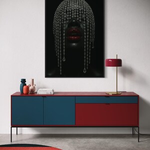Pode incluir: Uma impress&atilde;o de arte emoldurada com uma pessoa com uma cobertura de cabe&ccedil;a adornada com joias e l&aacute;bios vermelhos. Abaixo da obra de arte, h&aacute; um arm&aacute;rio moderno vermelho e azul com pernas pretas. Uma l&acirc;mpada vermelha e um tapete colorido completam a cena.
