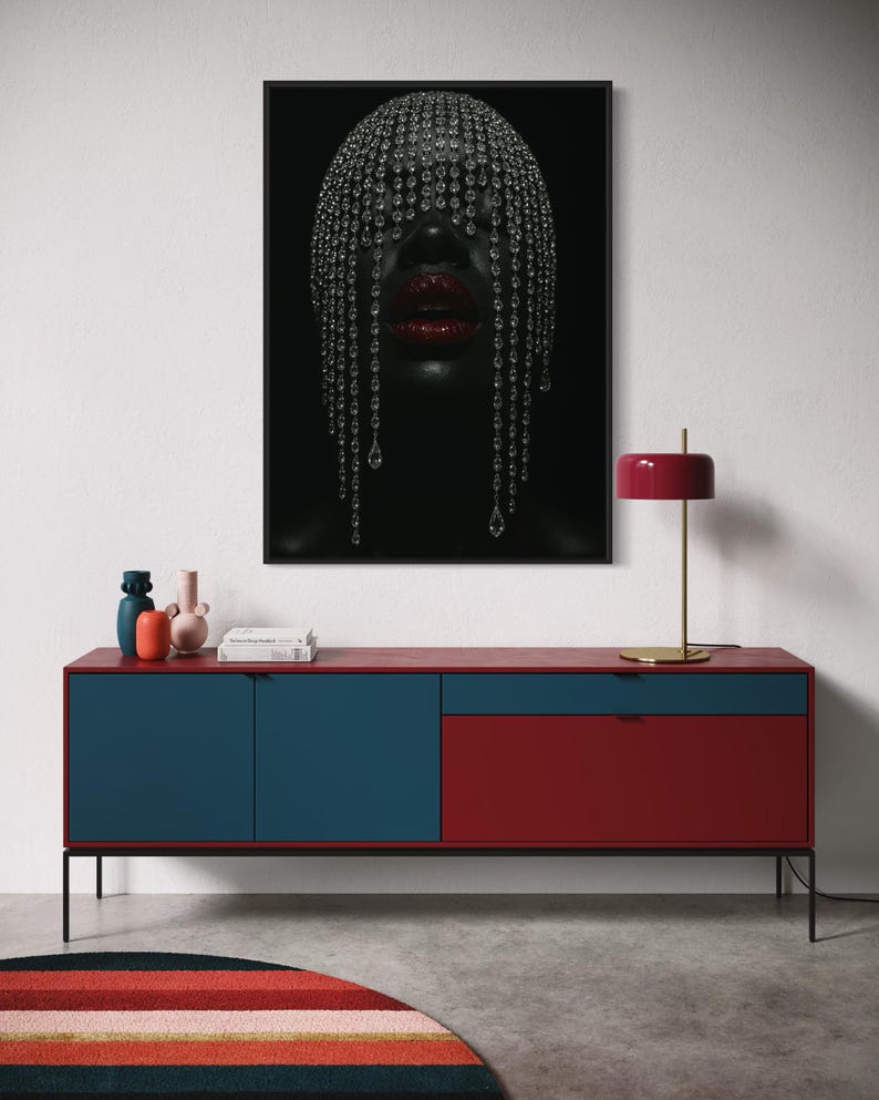 Pode incluir: Uma impress&atilde;o de arte emoldurada com uma pessoa com uma cobertura de cabe&ccedil;a adornada com joias e l&aacute;bios vermelhos. Abaixo da obra de arte, h&aacute; um arm&aacute;rio moderno vermelho e azul com pernas pretas. Uma l&acirc;mpada vermelha e um tapete colorido completam a cena.