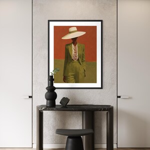 May include: A framed art print featuring a person in a green suit and a wide-brimmed hat, adorned with a pearl necklace. The background is divided into red and green sections. A black vase and decor sit on a dark table.