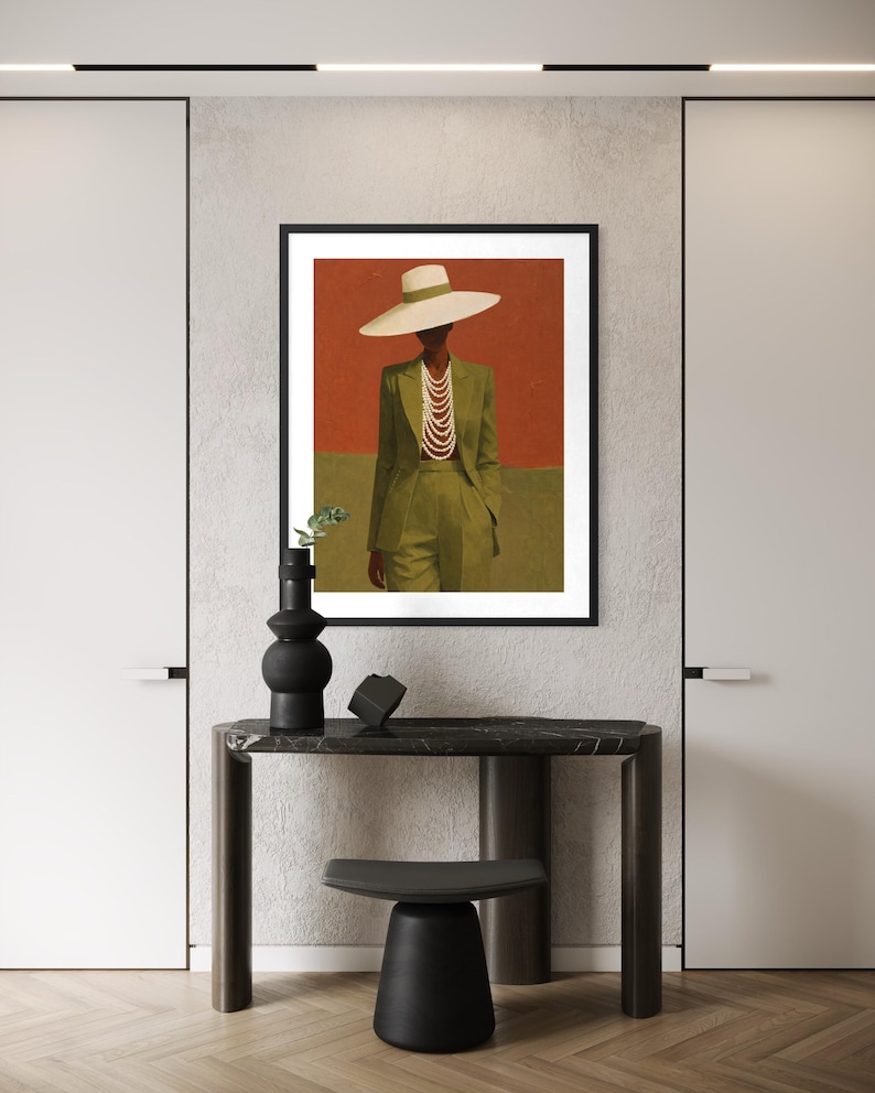 May include: A framed art print featuring a person in a green suit and a wide-brimmed hat, adorned with a pearl necklace. The background is divided into red and green sections. A black vase and decor sit on a dark table.