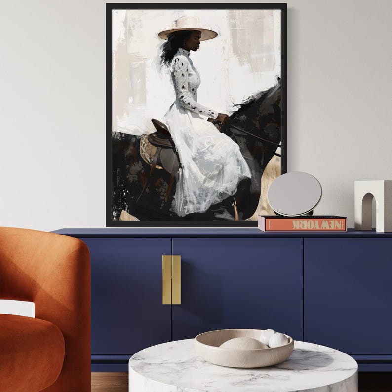 May include: A framed art print shows a woman in a white dress and hat riding a black horse. The artwork has a painterly style with neutral tones and a black frame. The woman is the focal point, with the horse's details visible.