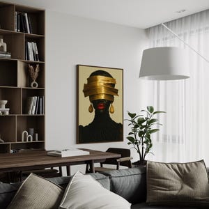 May include: An interior shot features a framed artwork with a portrait of a person with gold bands covering the eyes. The person is wearing a black top and gold earrings. The artwork hangs above a gray sofa with pillows, near a wooden table and bookshelf.