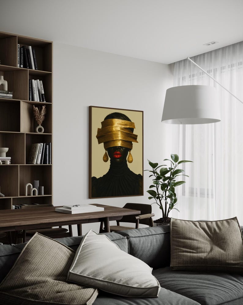 May include: An interior shot features a framed artwork with a portrait of a person with gold bands covering the eyes. The person is wearing a black top and gold earrings. The artwork hangs above a gray sofa with pillows, near a wooden table and bookshelf.