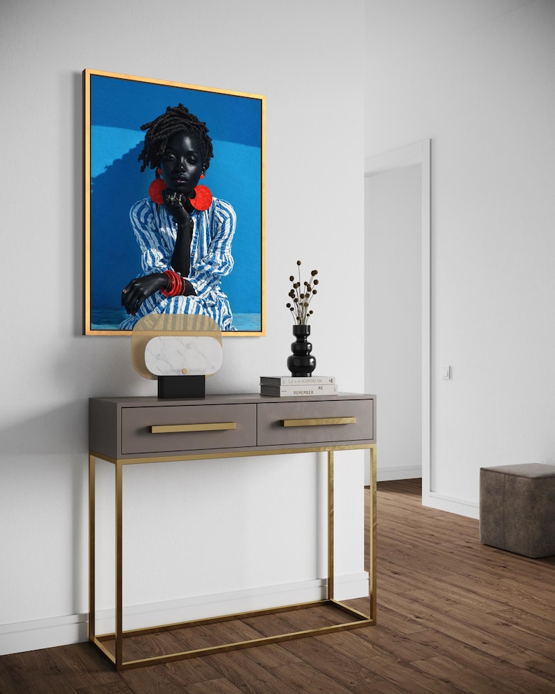 May include: A framed portrait of a person with dark skin wearing red jewellery and a blue and white striped outfit. The portrait hangs above a grey console table with gold legs and drawer pulls. A lamp and decorative vase are on the table.