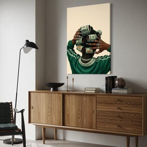 May include: A framed art print depicts a person with dollar bills rolled into curlers. The person wears a green and white striped shirt and a silver chain. The artwork hangs above a wooden cabinet with a black bowl and candles.