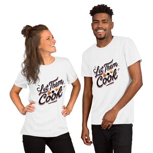 Peut inclure: Deux personnes portent des t-shirts blancs avec le texte "Let Them Cook" dans une police stylisée. Le motif comprend des illustrations d'articles liés à la cuisine. Les chemises sont à manches courtes et semblent être en coton.
