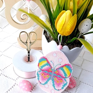 May include: An Easter-themed still life featuring a pink butterfly cross-stitch ornament with a colorful design. A spool of pink ribbon, gold scissors, and decorative eggs are arranged near a white egg-shaped planter with yellow tulips and greenery.