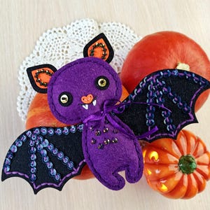 Op de afbeelding: Een paarse vilten vleermuis met zwarte paillettenvleugels en oranje accenten, geplaatst tussen kleine pompoenen. De vleermuis heeft goudkleurige ogen en een kleine oranje neus. Een wit kanten kleedje is zichtbaar op de achtergrond. Halloween-thema.