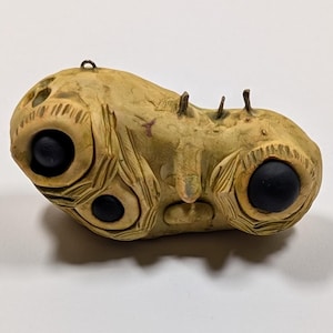 May include: A tan-colored, abstract sculpture with two large, black, circular eyes and a smaller eye below one. The sculpture has carved details and three small protrusions on top. A small metal loop is attached to the top.