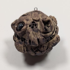 May include: A detailed, handcrafted ornament shaped like a grotesque, weathered face. The sculpture is primarily brown with dark accents, featuring a single, dark eye and intricate, textured details. A small metal loop is attached for hanging.