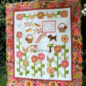 Puede incluir: Una colcha blanca con un borde floral rosa y marrón. La colcha presenta un diseño de patchwork con una variedad de patrones florales, incluyendo una casa, un arcoíris y un perro. El texto "There's No Place Like Home" está bordado en el centro de la colcha.