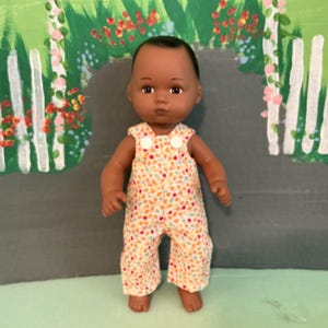 May include: A doll with dark skin and brown eyes wearing orange, yellow, and white patterned overalls. The overalls feature two white circular buttons at the top of the chest straps. The doll has a smooth, rounded head and a neutral facial expression.