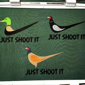 Może przedstawiać: Haftowany wzór na zielonym materiale z czarnym logo Nike swoosh i tekstem "Just Shoot It" powtórzonym trzy razy. Wzór przedstawia kaczkę, bażanta i złotego bażanta.