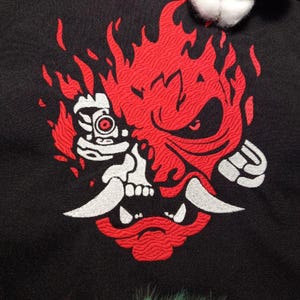 Puede incluir: Un parche bordado que representa una cara de demonio estilizada con llamas. El demonio tiene una calavera blanca con un ojo rojo y una boca roja con colmillos blancos.