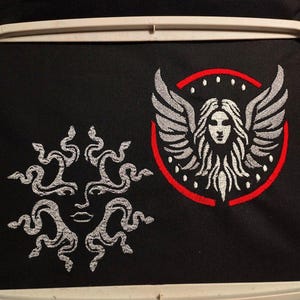 Puede incluir: Tela negra con dos diseños bordados. Uno es una cara solar con serpientes, el otro es una cara de mujer con alas dentro de un círculo rojo. Los diseños son en hilo blanco.