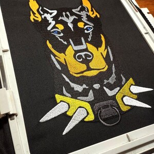 May include: Embroidered patch of a Doberman Pinscher dog with a spiked collar. The dog is black with yellow and white accents. The collar is yellow with white spikes.
