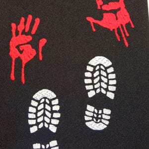 May include: Black fabric with two red handprints and three white footprints. The handprints are dripping with red paint. The footprints are in a line, with the largest footprint at the top.