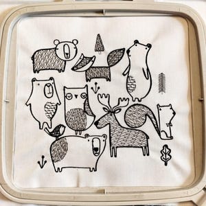 May include: Embroidered fabric featuring a whimsical forest scene. The design includes various animals such as bears, an owl, a fox, and a deer, all rendered in black thread on a white background. The embroidery is framed in a square hoop.