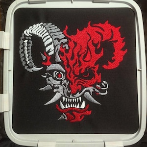 Pode incluir: Design bordado de um rosto de demônio estilizado com chifre de carneiro, chamas e dentes afiados. O design é principalmente vermelho, branco e cinza sobre um fundo preto, emoldurado por um bastidor de bordado branco. Para roupas ou acessórios.