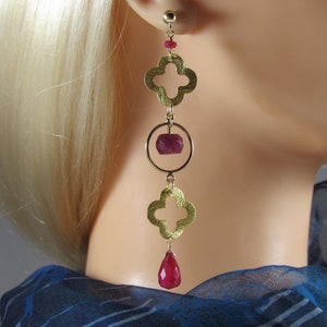 Pode incluir: Brincos dourados com design em forma de trevo e pedras preciosas vermelho rubi. Os brincos apresentam um design de gota com um aro redondo e um rubi em forma de lágrima na parte inferior. Usados por uma pessoa com cabelo loiro.