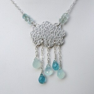 May include: A silver necklace featuring a cloud-shaped pendant with a delicate, openwork design. The necklace has small, light blue beads at the top and teardrop-shaped blue gemstones hanging below, creating a rain-like effect. The chain is also silver.