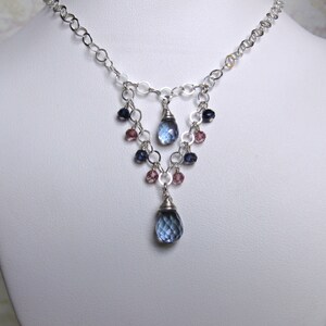 May include: A silver chain necklace with a teardrop-shaped blue gemstone pendant. The pendant is surrounded by smaller blue and pink gemstones.